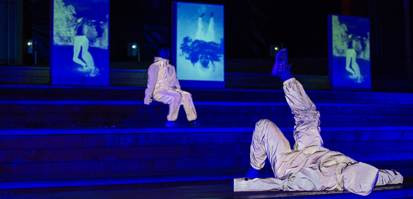 Photo in blue light of two people sitting and laying on stairs wearing white suits.