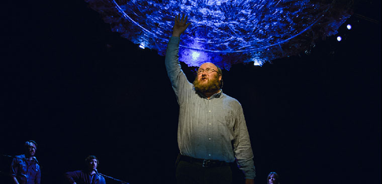 Photo from a past Artistic Fraud presentation of a man on stage standing on a chair reaching towards the ceiling.