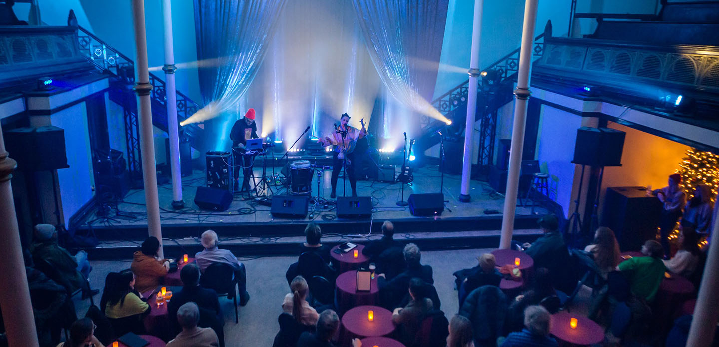 Photo of an indoor event with 2 musicians performing on a stage and audience seated at tables.