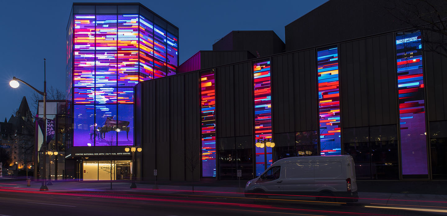 Photo of Canada’s National Art Centre taken from the street at night with lights on all windows on display lit up in full colour. 